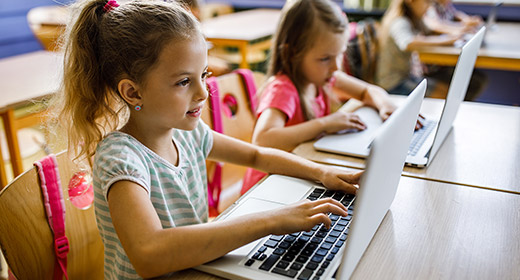 Two young girls sitting at desks in a classroom taking notes on laptop computers