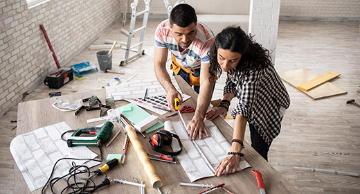 Middle age couple standing in a room that is being renovated the couple is looking at blueprints and measuring a table