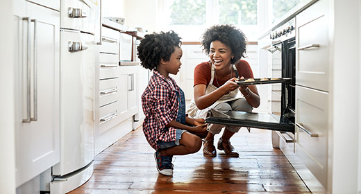 Mom with her young daughter baking and putting cookies in an oven in the kitchen