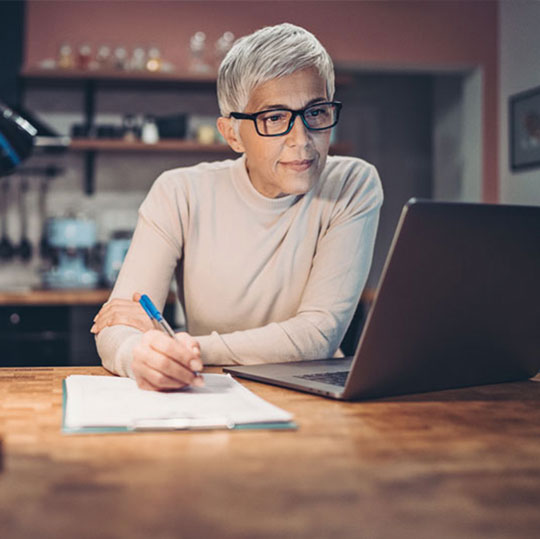 Older woman writing in a notebook while looking at her computer