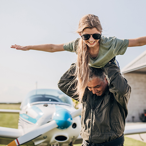 father holding his young daughter above his head in front of an airplane_480x480
