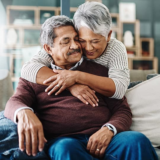 older man sitting on a couch while his wift hugs him while standing behind the couch