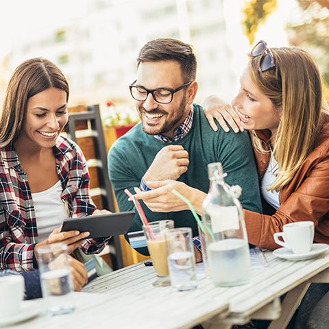 three friends sitting on restaurant patio looking at a tablet together 480x480