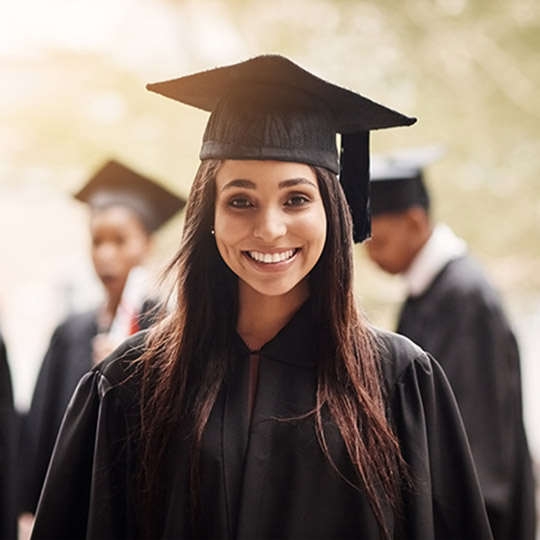 Girl in graduation cap and gown smiling at the camera while other graduates mingle behind her
