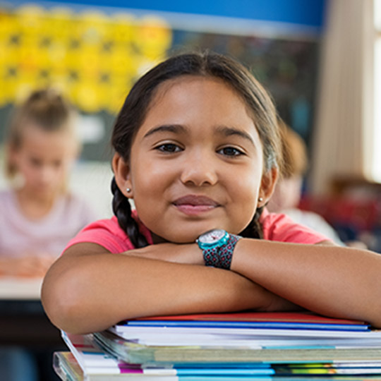 Young girl in a classroom leaning on a pile of books on her desk