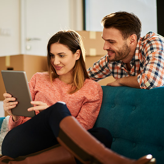 Younger couple looking at an iPad on a couch sorrounded by moving boxes in their new home_1