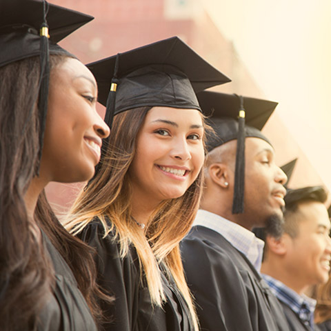 College graduates in cap and gown standing in a line while one of the girls smiles at the camera