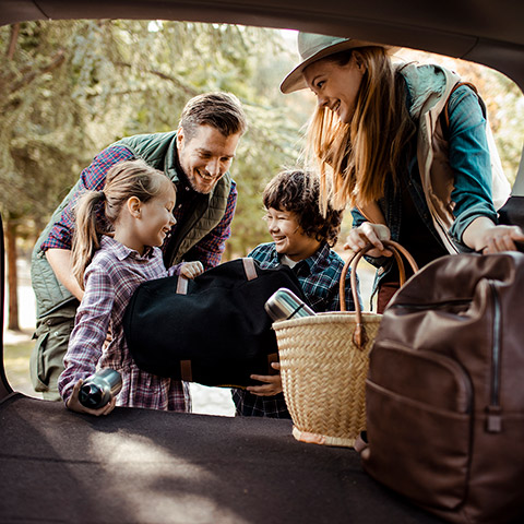 Family of four two kids two parents packing bags into the back of their van for vacation while talking and laughing