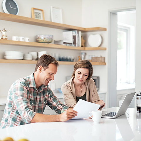 Man and woman sitting at their kitchen island discussing finances and utilizing paper and a laptop