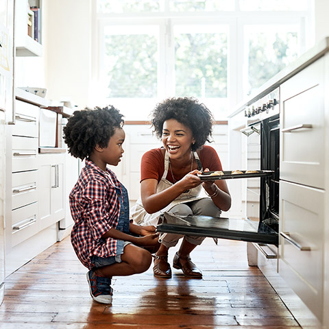 Mom with her young daughter baking and putting cookies in an oven in the kitchen