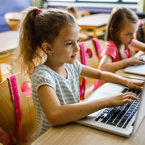 Two young girls sitting at desks in a classroom taking notes on laptop computers