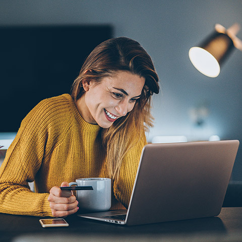 Woman smiling while online shopping at laptop on a desk and hold a coffee mug and credit card