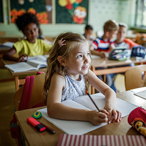 Young girl sitting at a desk in school listening to a teacher while taking notes with a pencil